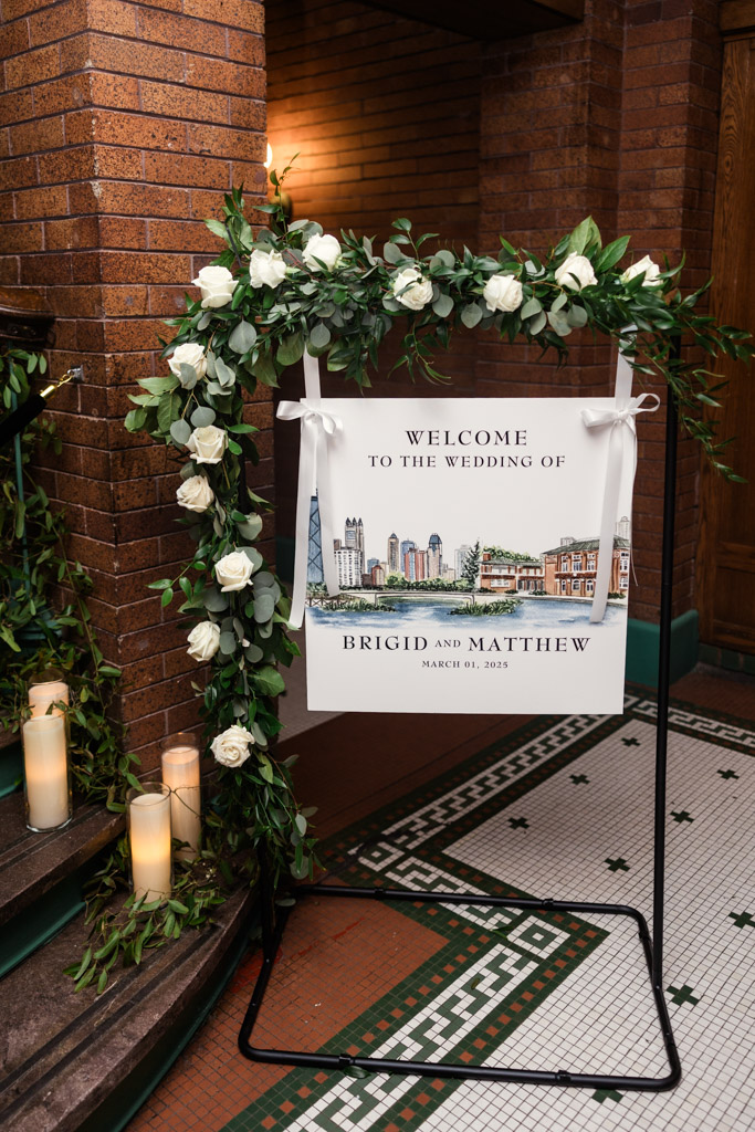 Wedding welcome sign with florals, candles, and an illustrated city skyline for wedding reception at Cafe Brauer