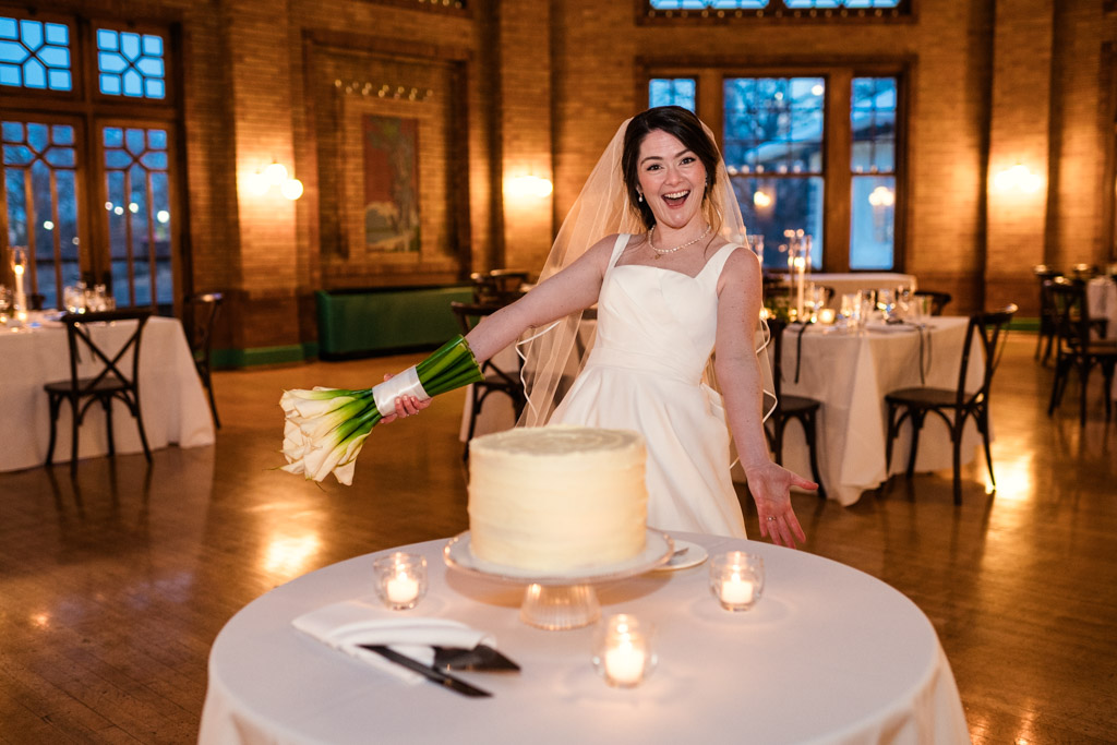 Bride in a white dress smiles, holding her bouquet next to the wedding cake she made for her wedding reception at Cafe Brauer
