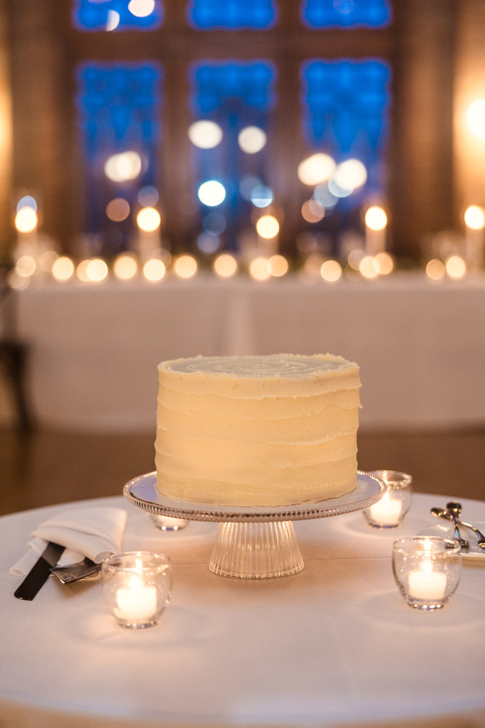 White frosted wedding cake on a stand, surrounded by candles, baked by the bride for Cafe Brauer wedding reception