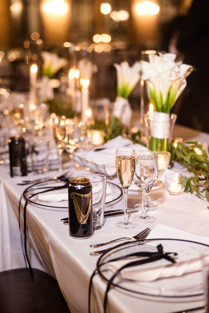 Guest table set with white linens, floral centerpieces, candles, glassware, and a can of Guinness for Cafe Brauer wedding reception