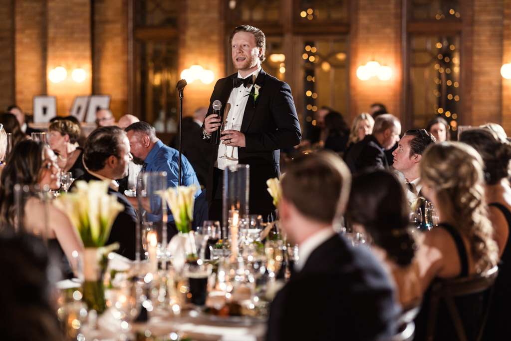 Best Man in a tuxedo gives a speech at a wedding reception in Cafe Brauer, surrounded by seated guests at decorated tables