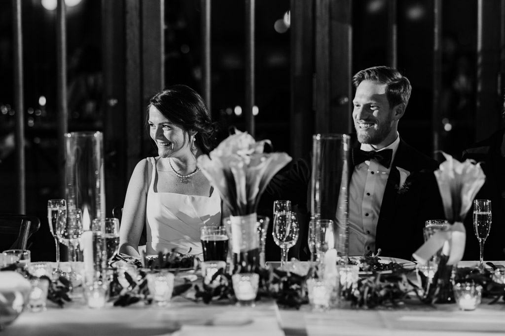 Black and white photo of bride and groom sitting and smiling at the head table during their wedding reception at Cafe Brauer