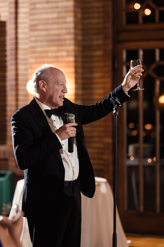 Father of the bride in a tuxedo raises a wine glass and speaks into a microphone at Cafe Brauer wedding reception