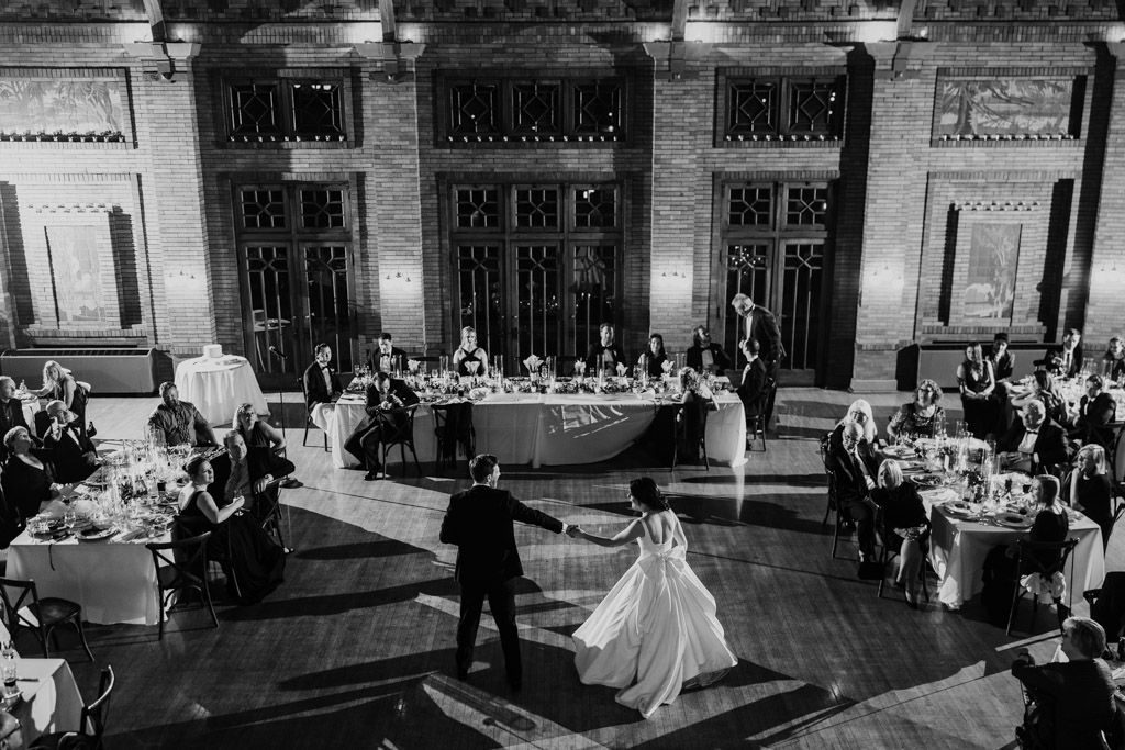 Black and white photo of bride and groom dancing in the center of the elegant Cafe Brauer hall as reception guests watch from decorated tables