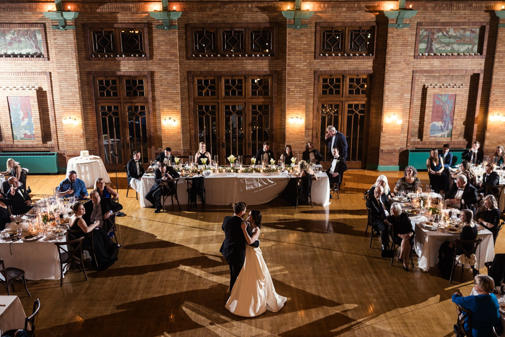 Newlyweds share a first dance in the grand hall of Cafe Brauer, surrounded by seated wedding guests