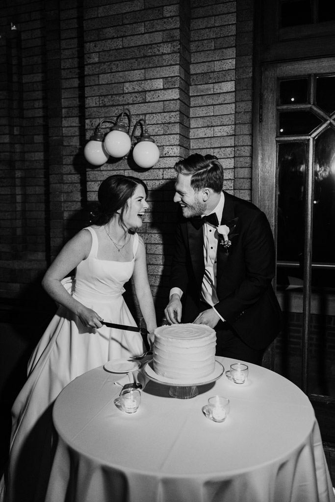 Black and white photo of bride and groom smiling at each other as they get ready to cut their wedding cake at their Cafe Brauer wedding reception