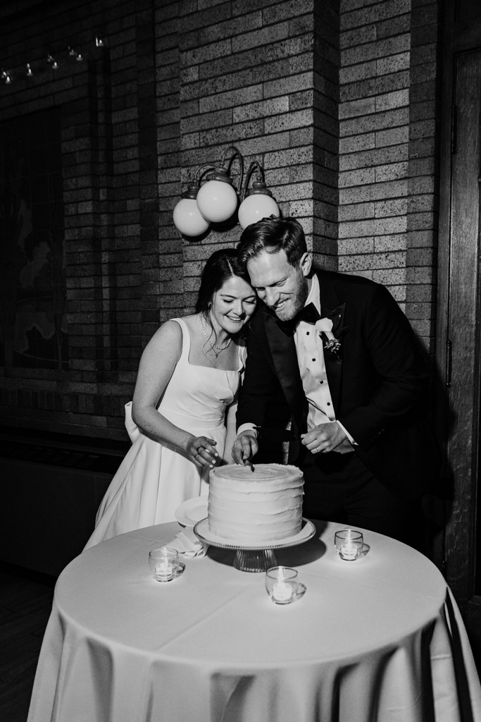 Black and white photo of bride and groom smiling as they cut their wedding cake together during their Cafe Brauer wedding reception