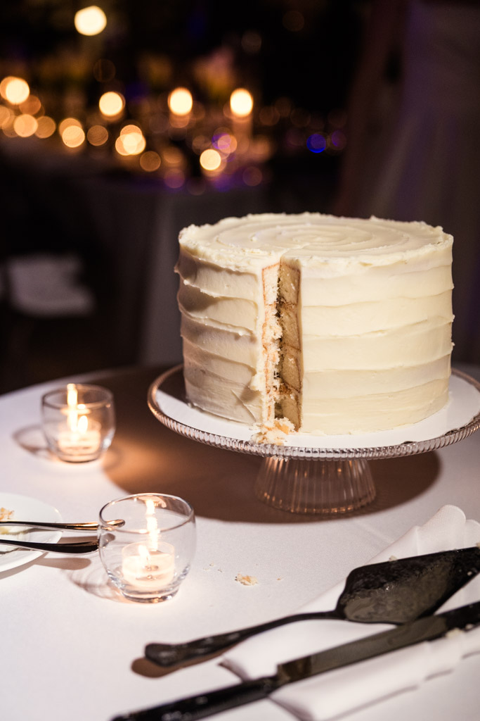 White frosted wedding cake with a slice missing sits on a glass stand beside candles on a table for wedding reception at Cafe Brauer