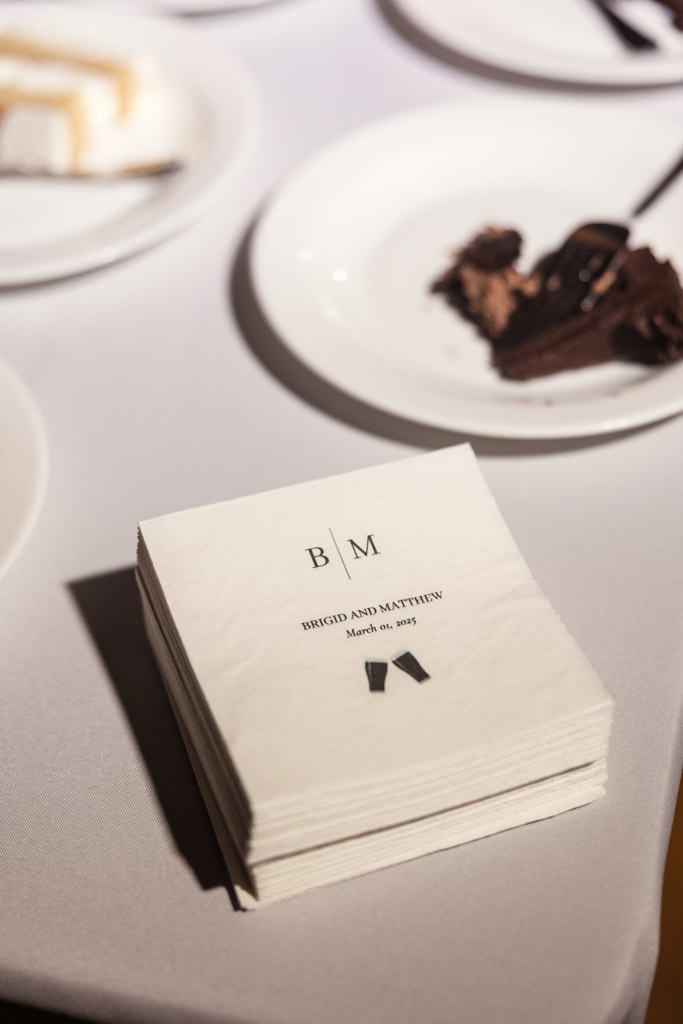 Stack of custom napkins with newlyweds' initials and plates of cake on a white tablecloth for Cafe Brauer wedding reception
