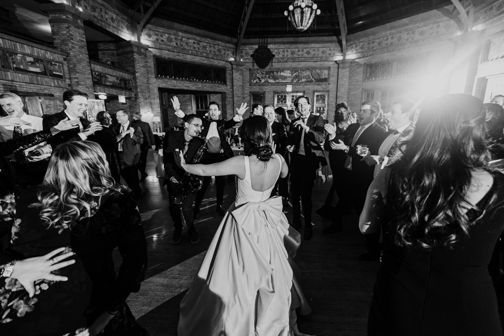 Black and white of bride in the center of a lively group of guests on the dance floor at her Cafe Brauer wedding reception