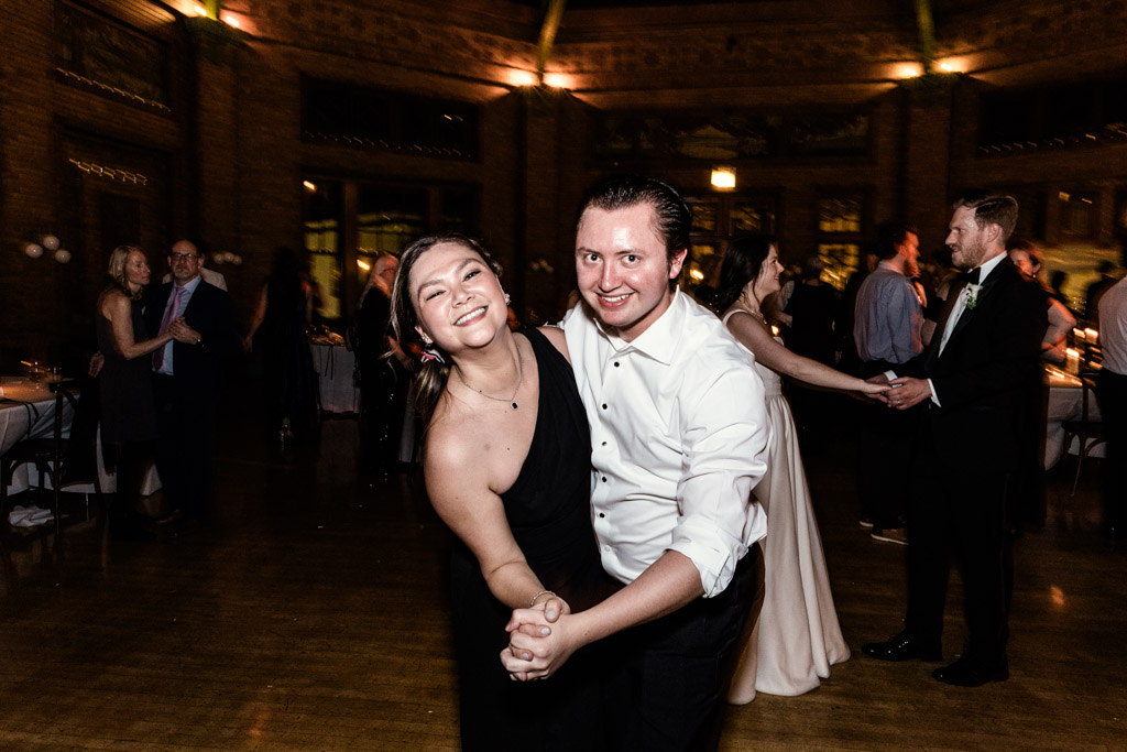 A smiling couple dances together during a wedding reception at Cafe Brauer, surrounded by other guests on a dimly lit dance floor