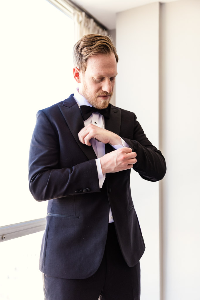 Groom in a black tuxedo adjusts his cufflink near a window with natural light inside his apartment