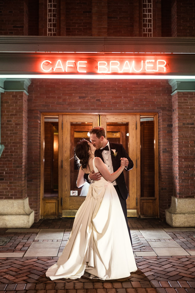 Newlyweds kiss outside their Cafe Brauer wedding reception at night, illuminated by the glow of the venue's red neon sign