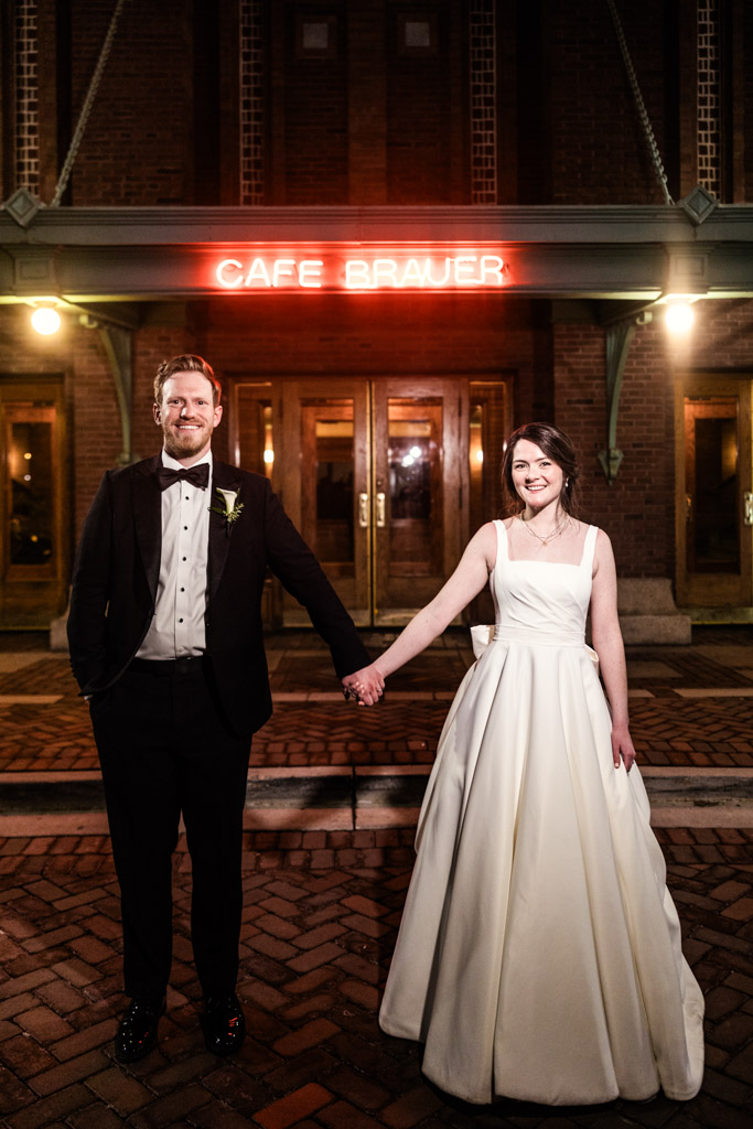 Portrait of bride and groom holding hands, standing outside Cafe Brauer at night during their wedding reception