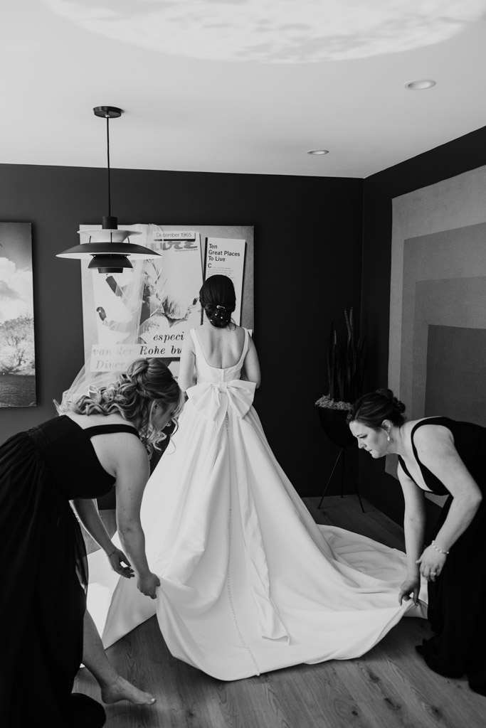 Black and white photo of two bridesmaids adjusting the train of bride's dress inside the bride's apartment before her Cafe Brauer wedding celebration