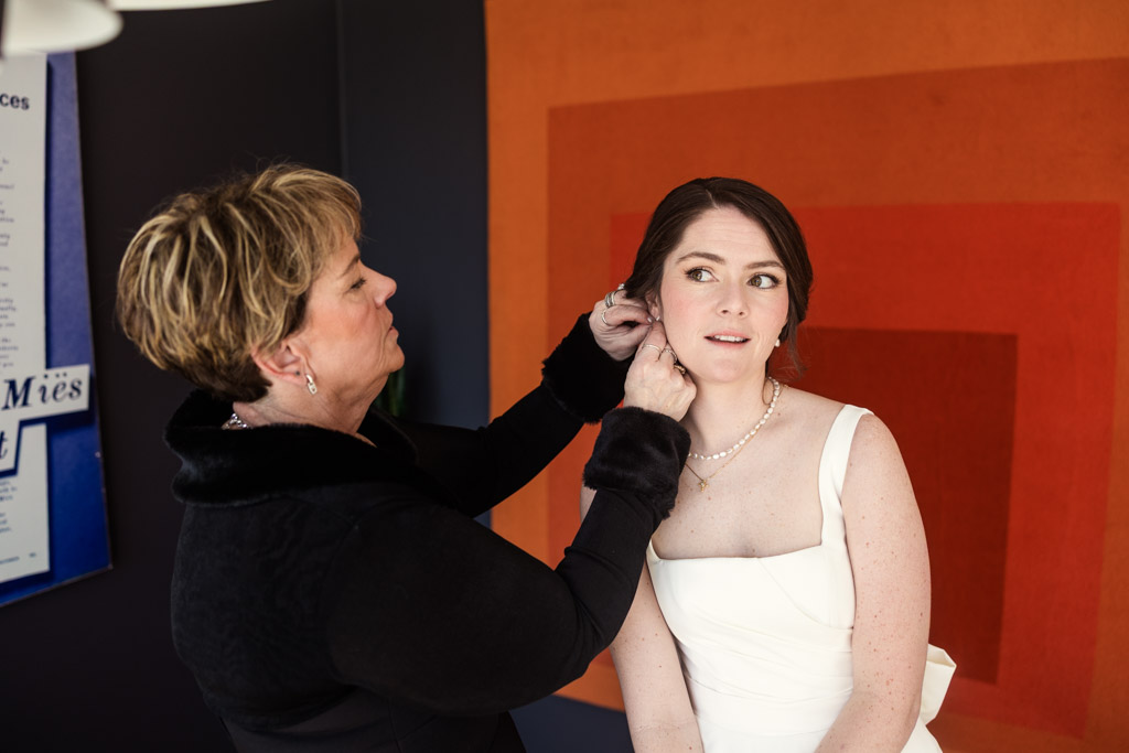 Bride's mother helps bride put on earrings as the bride sits in a white dress before her spring Chicago wedding celebration
