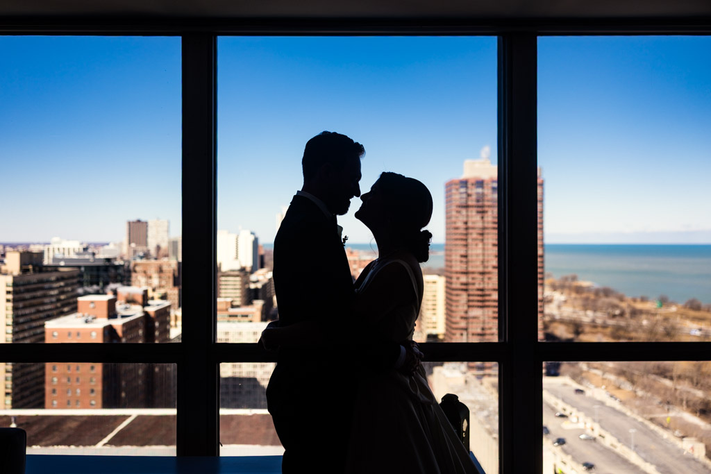 Silhouetted couple embraces by a large window inside their Lincoln Park apartment, overlooking Chicago and Lake Michigan