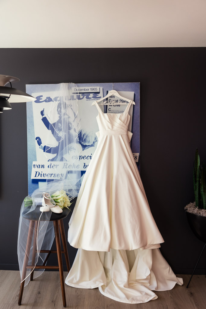 Bride's dress, veil, shoes, and bouquet displayed against a dark wall with artwork inside the couple's apartment before their spring Chicago wedding celebration