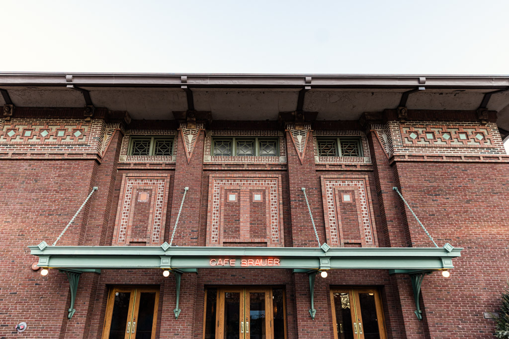 Cafe Brauer’s brick facade features geometric designs, a green awning, and a red neon Cafe Brauer sign glowing above its wooden doors