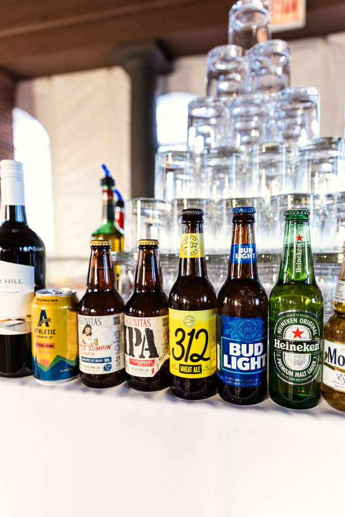 Assorted beer and beverage bottles and cans lined up on a counter for Cafe Brauer wedding reception, with stacked glasses in the background