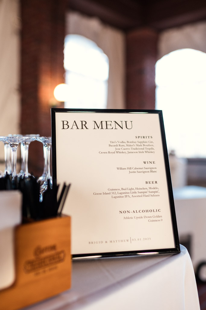 Framed bar menu listing spirits, wine, beer, and non-alcoholic drinks on a white-draped table at Cafe Brauer wedding reception
