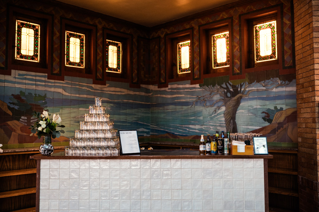 Bar with stacked glasses, bottles, and a mural of a tree and river beneath five stained glass windows in wedding reception space at Cafe Brauer in Chicago