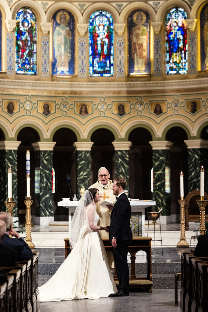 Bride and groom stand at the altar with a priest during their wedding ceremony inside St. Clement Church in Chicago