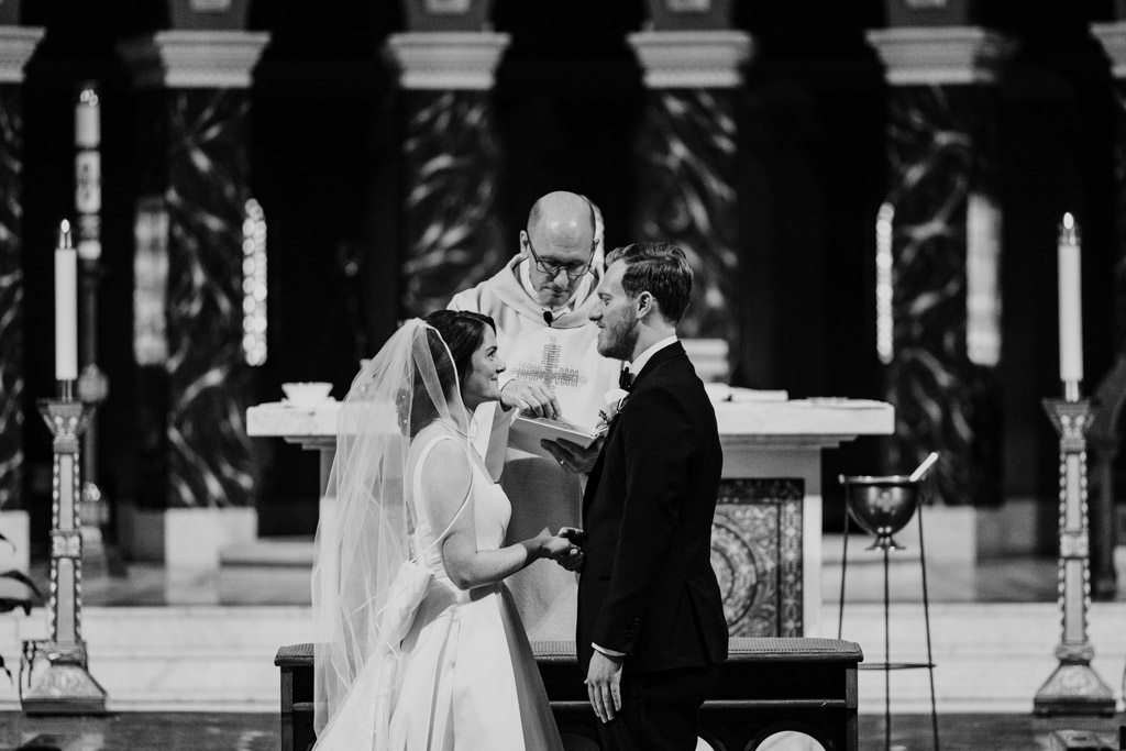 Black and white photo of bride and groom exchanging vows in front of the altar during their wedding ceremony at St. Clement Church in Chicago