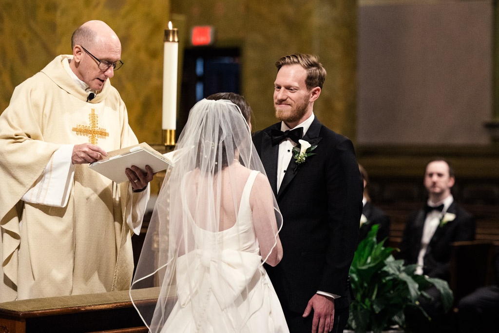 A bride and groom stand at the altar with a priest during their wedding ceremony at St. Clement Church in Chicago