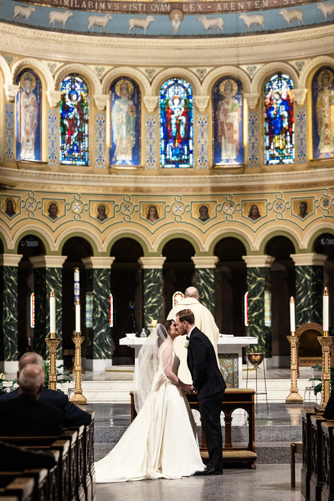 Bride and groom kiss at the altar of St. Clement Church in Chicago, with stained glass windows behind them