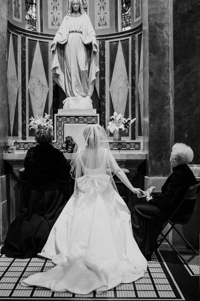 A bride kneels at an altar for St. Mary with her mother and grandmother seated beside her during wedding ceremony at St. Clement Church in Chicago