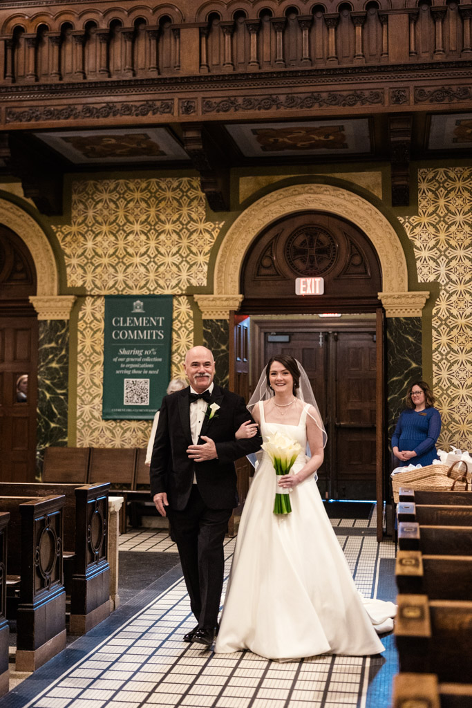 A bride in a white gown walks down the aisle arm in arm with her father during St. Clement Church in Chicago wedding ceremony