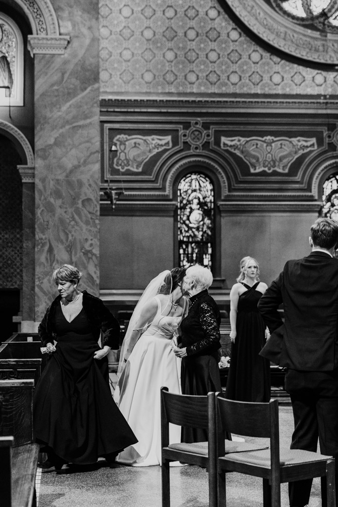 Black and white photo of bride kissing her grandmother inside St. Clement Church in Chicago, surrounded by guests in formal attire