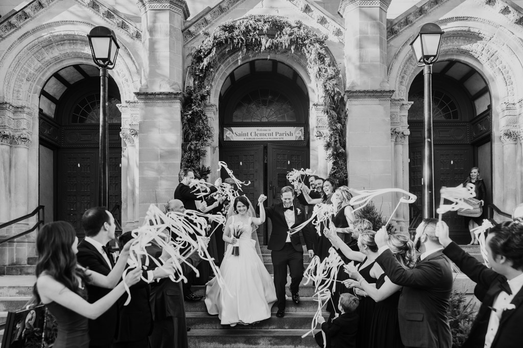 Black and white photo of newlyweds exiting St. Clement Church in Chicago as guests cheer and wave streamers on the steps outside