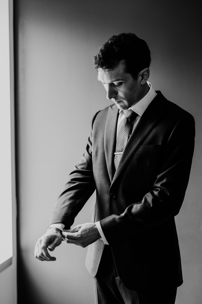 Groom adjusts his cuff while standing near a window atThe Godfrey Hotel in downtown Chicago illuminated by natural light