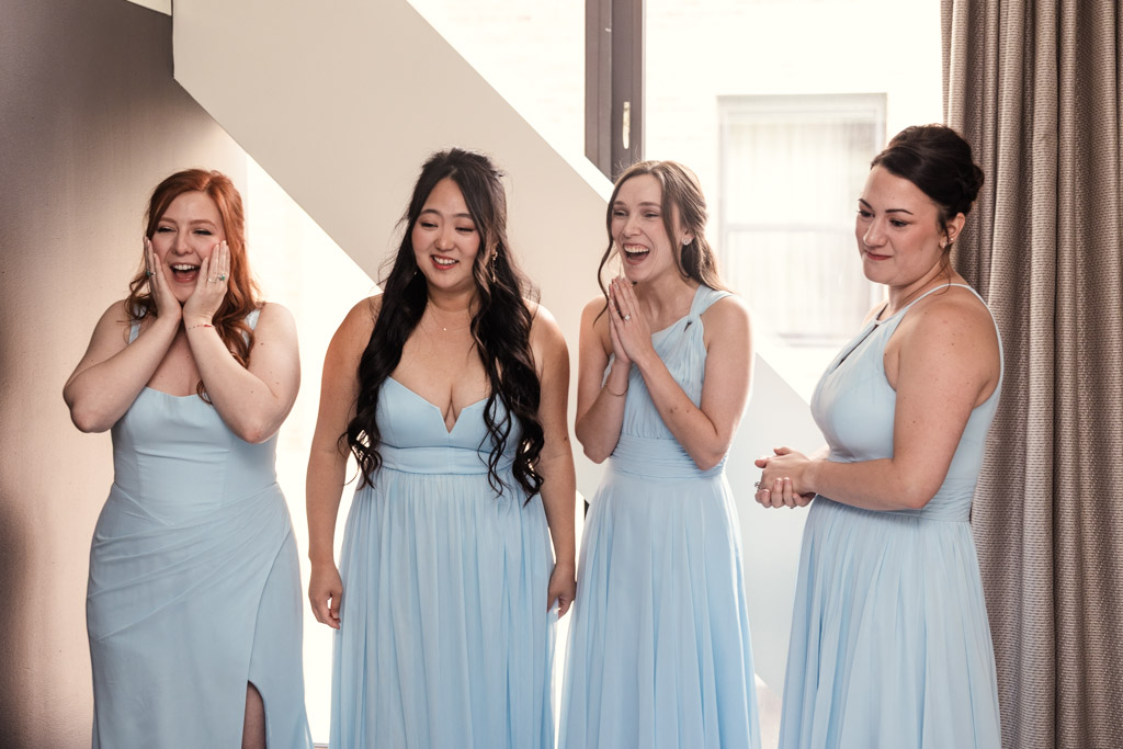Four bridesmaids in light blue dresses smile and react to seeing the bride in her gown after getting ready at The Godfrey Hotel in downtown Chicago