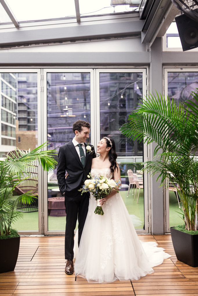 Bride and groom smile at each other indoors at The Godfrey Hotel in downtown Chicago, surrounded by lush green plants