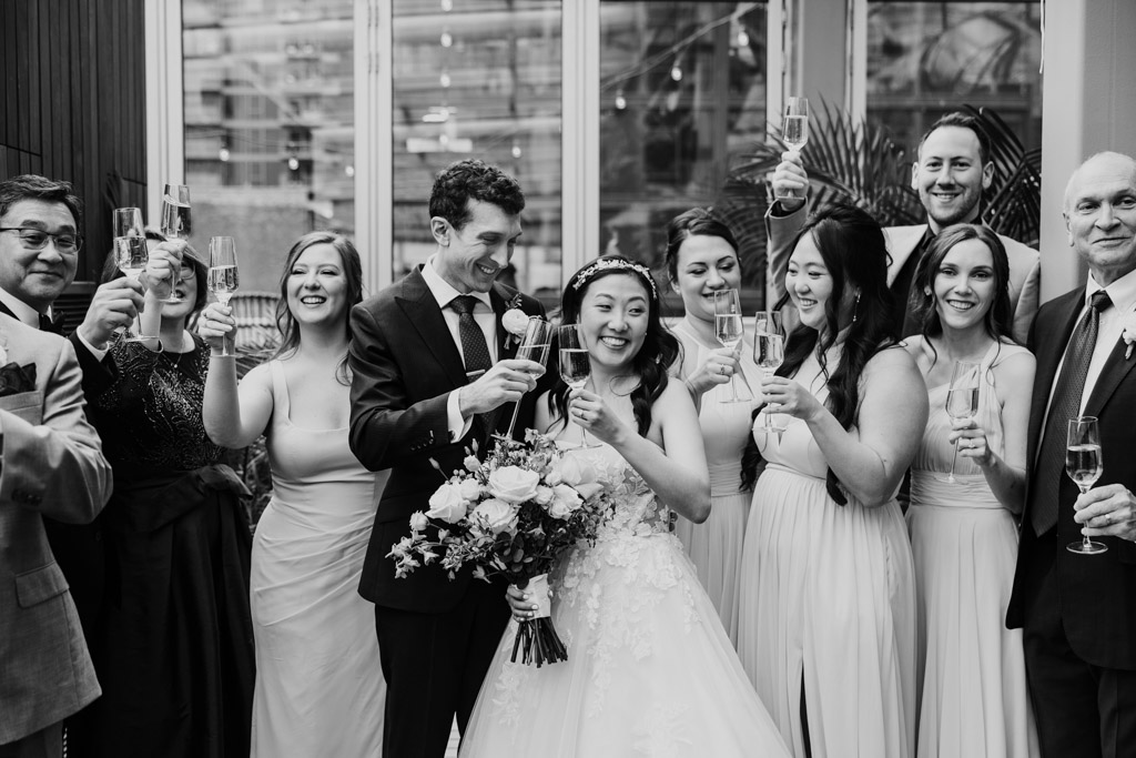Black and white photo of bride and groom smiling and toasting with their wedding party and families at Chicago Winery