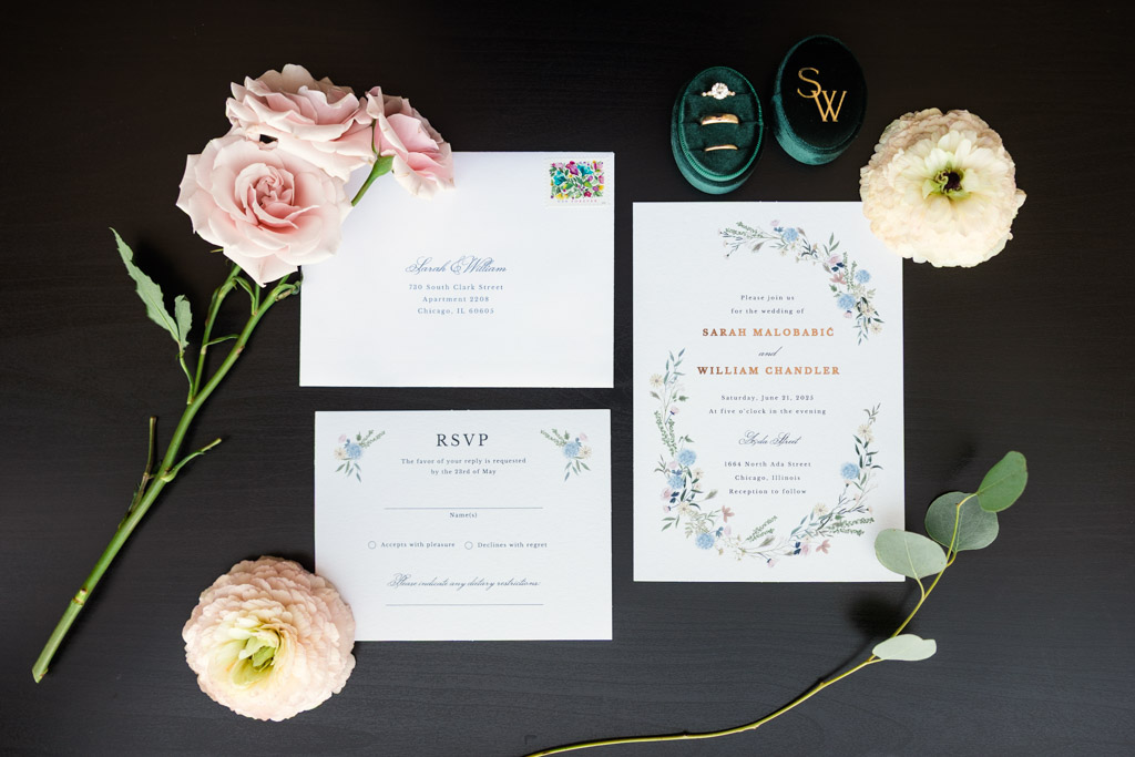 Wedding invitations, RSVP card, pink flowers, and a green ring box with gold rings set on a dark table for your Ada Street Chicago wedding celebration