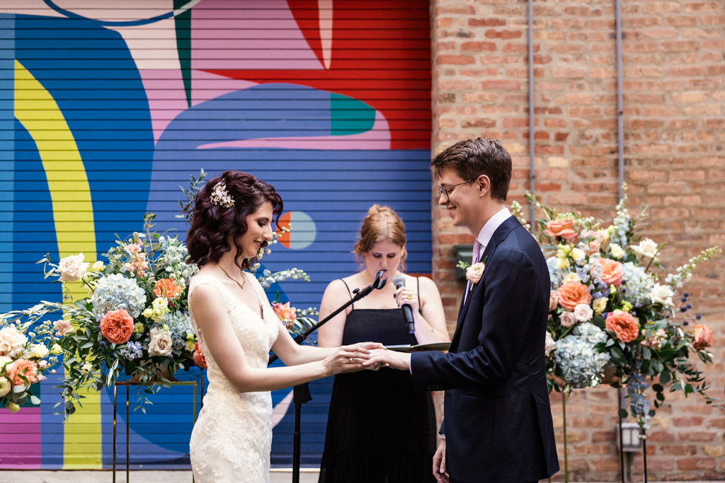 Bride places ring on groom's finger during their wedding ceremony at Ada Street Chicago, surrounded by colorful floral arrangements and a vibrant mural