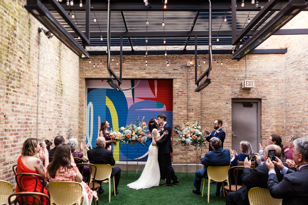 Newlyweds kiss during their indoor Ada Street Chicago wedding ceremony, surrounded by guests and colorful floral arrangements