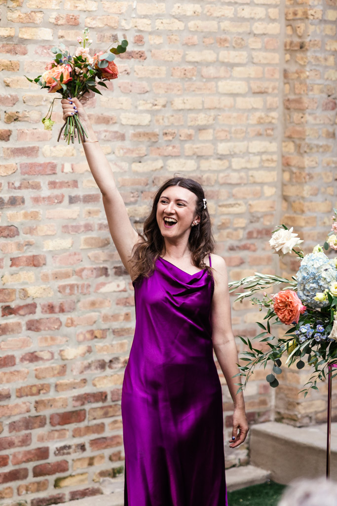 Maid of Honor in a purple dress smiles and raises a bouquet in the air, standing by a brick wall during Ada Street Chicago wedding reception
