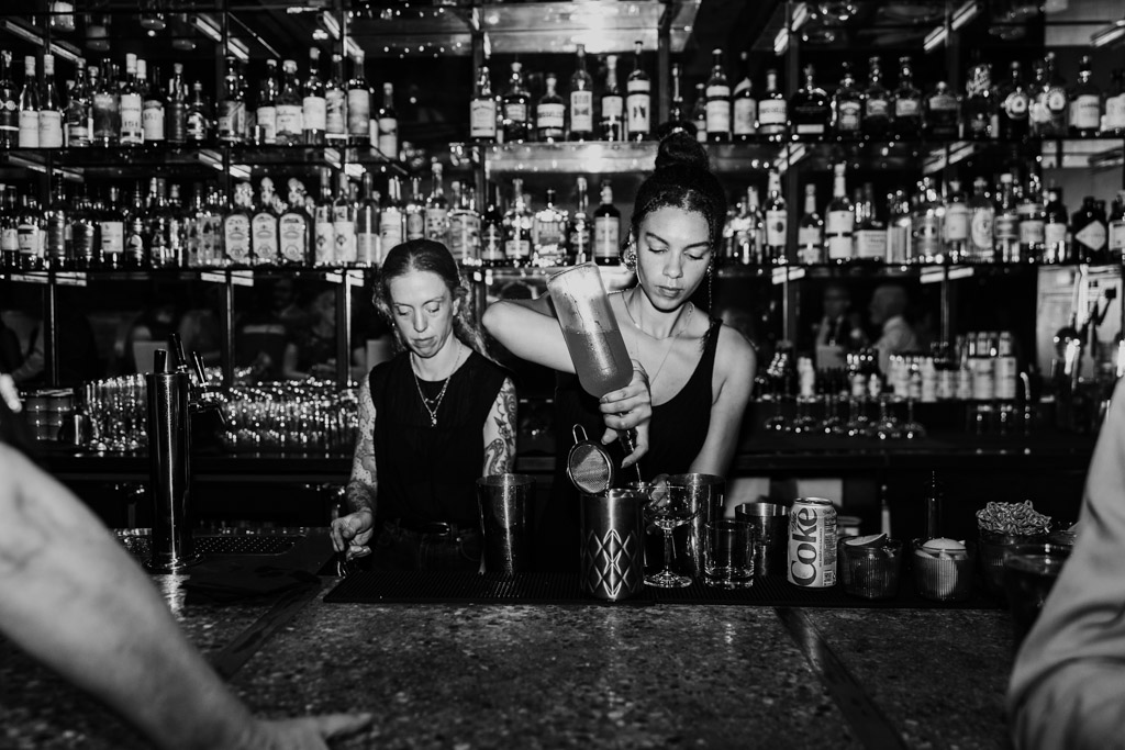 Black and white photo of two bartenders mixing drinks behind a busy, well-stocked bar at Ada Street Chicago