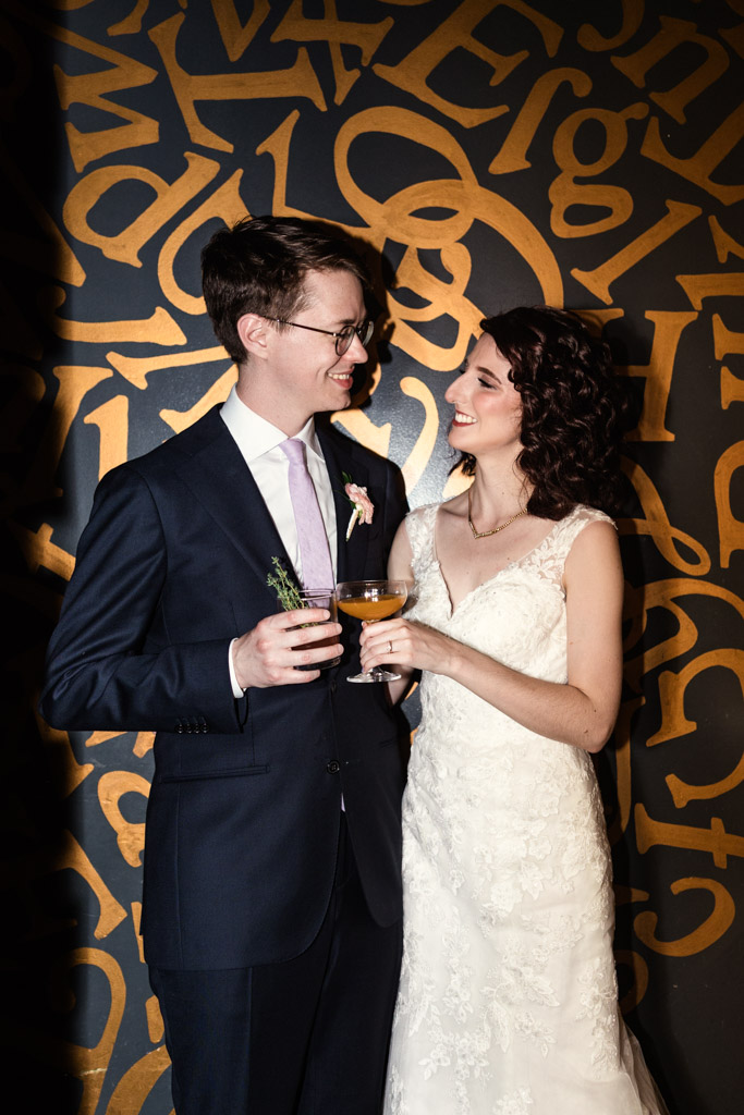 A bride and groom smile at each other, holding drinks, in front of a gold and black lettered wall at Ada Street Chicago