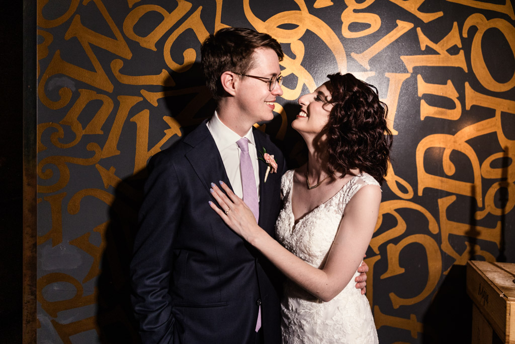 A couple in wedding attire smiles and embraces in front of a wall with gold letters on Ada Street Chicago.