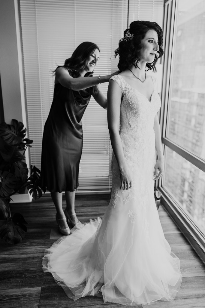 Black and white photo of woman helping bride with her dress as they stand by a window before wedding celebration at Ada Street Chicago