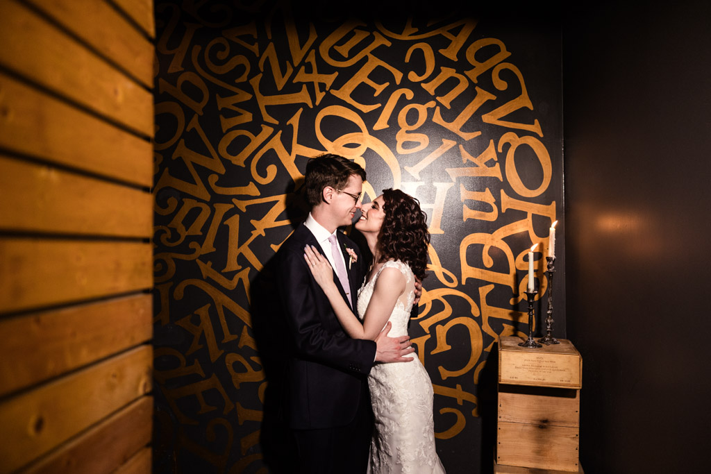 Bride and groom embrace and smile in front of a wall with gold letters and a wooden candle holder at Ada Street Chicago