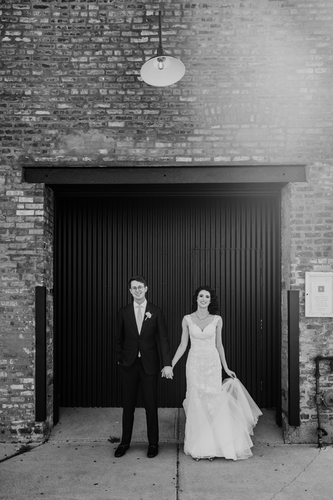 Black and white portrait of bride and groom holding hands, standing in front of a brick wall and large metal door at Ada Street Chicago