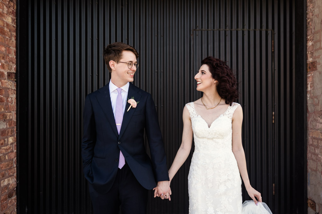 Bride and groom holding hands, smiling at each other in front of a black, striped metal wall at Ada Street Chicago