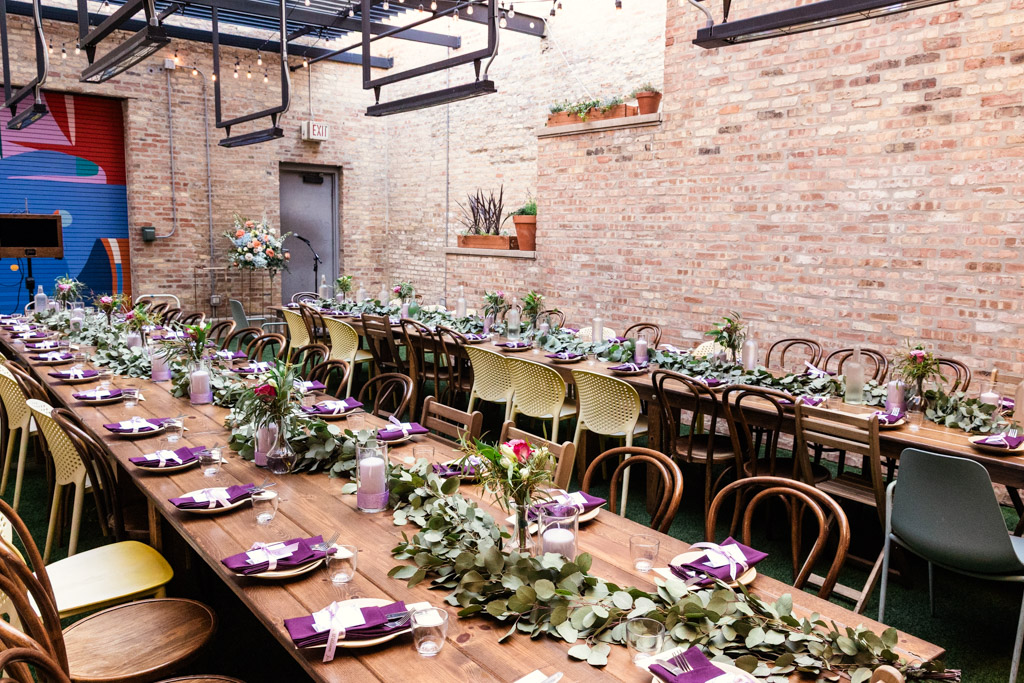 Long wooden tables set for wedding reception with greenery, purple napkins in a brick-walled room at Ada Street Chicago
